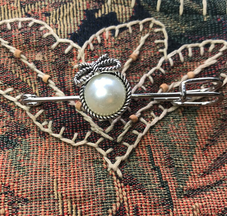 May include: A silver-toned brooch with a large, round, faux pearl center. The pearl is surrounded by a twisted rope design and topped with a bow. The brooch has a pin closure and is set against a patterned fabric.