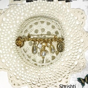 May include: A gold-toned vintage brooch with a safety pin closure. The brooch features a chain design with dangling faux pearls, crystals, and coin-shaped charms. The brooch is set against a white, lacy background.