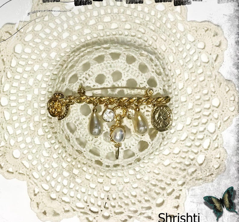 May include: A gold-toned vintage brooch with a safety pin closure. The brooch features a chain design with dangling faux pearls, crystals, and coin-shaped charms. The brooch is set against a white, lacy background.