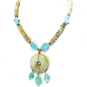 May include: A handcrafted necklace featuring a large, pale green circular pendant with gold wire accents and three dangling aqua-colored teardrop-shaped beads. The necklace includes light green and beige beads, and aqua-colored glass beads.