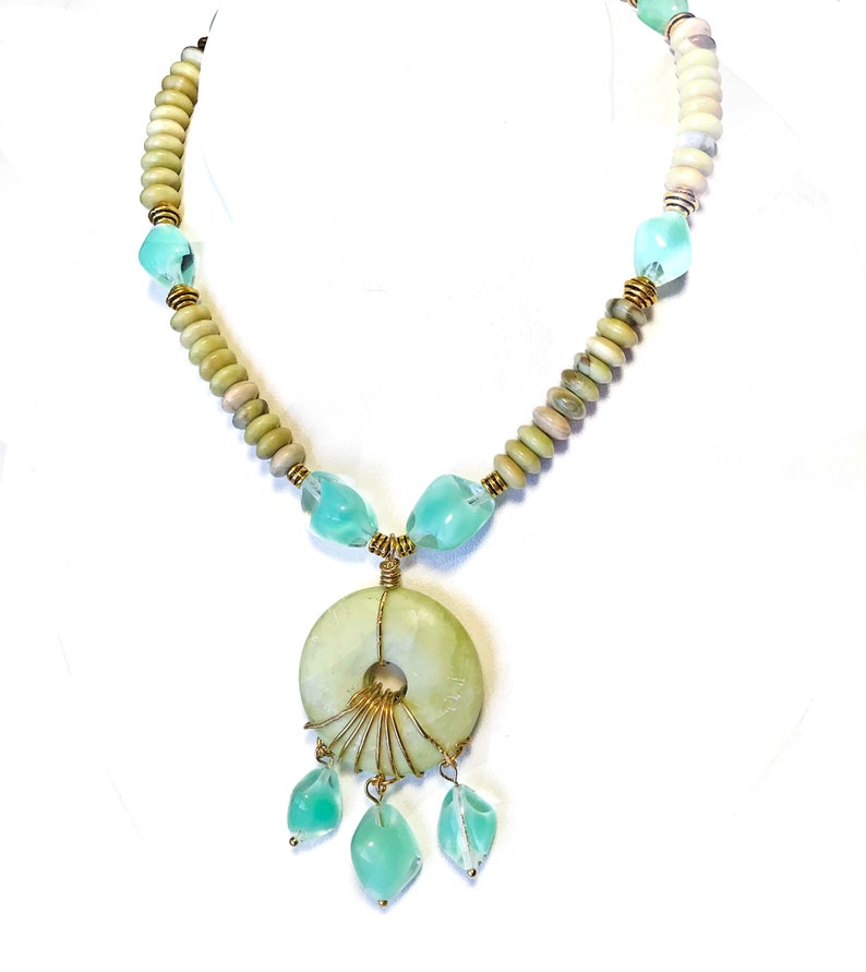 May include: A handcrafted necklace featuring a large, pale green circular pendant with gold wire accents and three dangling aqua-colored teardrop-shaped beads. The necklace includes light green and beige beads, and aqua-colored glass beads.
