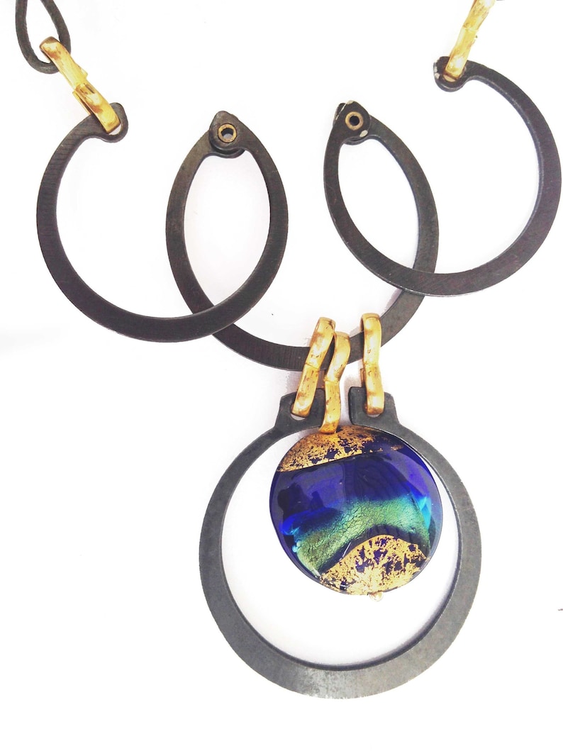 May include: A handcrafted necklace featuring a dark metal chain with interlocking oval and circular links. The pendant showcases a blue and green glass orb with gold accents, suspended within a circular frame.