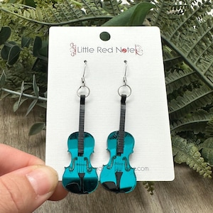 May include: A pair of teal violin-shaped earrings with black details, hanging from silver hooks. The earrings are displayed against a white card with the text "Little Red Note" and a background of green foliage.