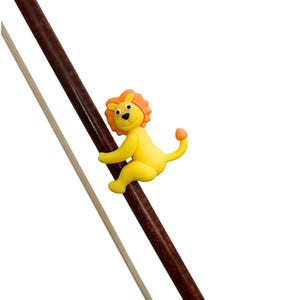 Può includere: Una statuetta a forma di leone giallo con criniera e coda arancione si aggrappa a un bastoncino di legno marrone scuro. Un bastoncino di legno sottile e di colore chiaro è posizionato parallelamente al bastoncino marrone. Lo sfondo è bianco.