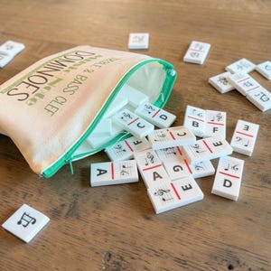 Könnte beinhalten: Ein Musik-Domino-Set mit einer Canvas-Tasche. Die Dominosteine sind weiß mit Noten und Buchstaben. Die Tasche ist beige mit einem grünen Reißverschluss und den Worten "DOMINOES TREBLE & BASS CLEF".