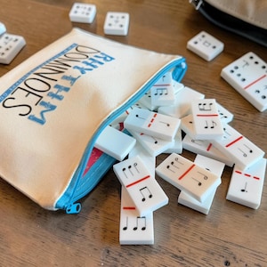 May include: A canvas pouch with the text "Rhythm Dominoes" spills out white dominoes with musical notes and red lines. Some dominoes are scattered on a wooden surface. A blue zipper secures the pouch.