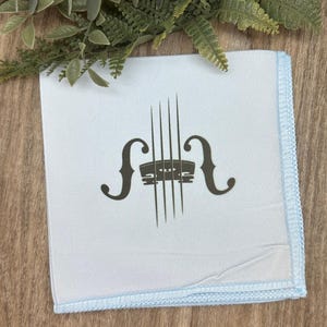 May include: A light blue handkerchief with a dark gray violin graphic. The handkerchief has a light blue border and is displayed on a wooden surface. The design features the shape of a violin's bridge and strings.