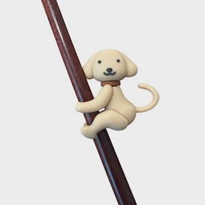 May include: A cream-colored dog figurine with a brown collar is attached to a thin, dark brown wooden rod. The dog has floppy ears, a small black nose, and a curved tail, appearing to climb or cling to the side of the straight wooden stick.