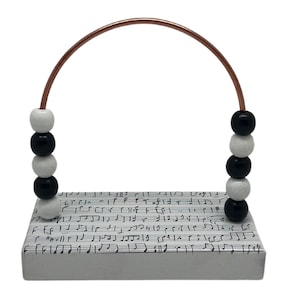 May include: A white wooden base with black musical notes printed on it. A copper wire arch is attached to the base with black and white beads strung on the wire.