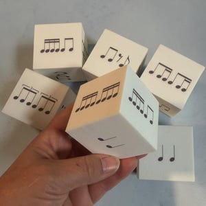 May include: A collection of white foam cubes featuring black printed musical notation symbols on their faces. Each cube displays various rhythmic patterns, including beamed eighth notes, sixteenth notes, and triplets, designed for music education.