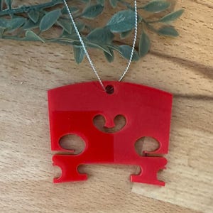 Puede incluir: Un adorno de puente de violín rojo brillante con una cuerda plateada para colgar. El adorno tiene una forma de puente clásica con detalles recortados. El fondo es una superficie de madera con una ramita de vegetación.