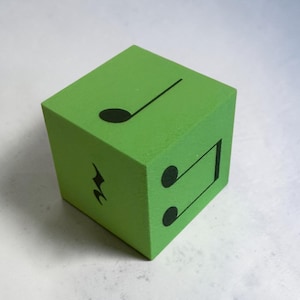 Puede incluir: Un cubo de madera de color verde lima brillante con símbolos de notación musical negros impresos en sus caras visibles. La cara superior muestra una negra, mientras que las caras laterales presentan un silencio de negra y dos corcheas unidas.
