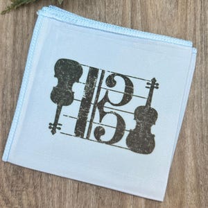 Puede incluir: Un pañuelo azul claro con un tema musical. El pañuelo presenta un diseño negro desgastado de una clave de sol y dos violines. Los bordes están ribeteados con costuras azul claro.