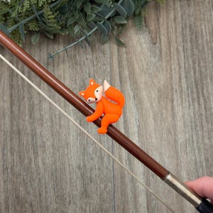 Puede incluir: Un arco de violín marrón con una pequeña figura de zorro naranja adjunta. El zorro tiene detalles blancos y está posado en el arco. El arco es sostenido por una mano, y el fondo es una superficie de madera con vegetación.