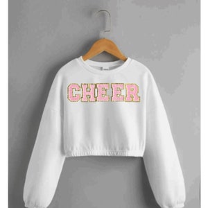 May include: White cropped sweatshirt with long sleeves and a pink and gold "CHEER" graphic.