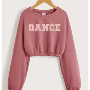 May include: A pink cropped sweatshirt with long sleeves and a gold and pink "DANCE" graphic on the front.