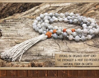 May include: Gray gemstone mala necklace with a white tassel, strung on organic hemp cord. The text "STRUNG ON ORGANIC HEMP CORD: THE STRONGEST & MOST ECO-FRIENDLY NATURAL FIBER ON EARTH." is printed on a brown label.