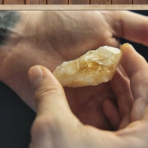 May include: A hand holding a light brown, translucent crystal with a pointed end. The crystal has a natural, rough texture.