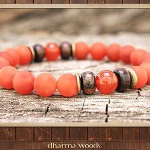 May include: A stretch bracelet featuring matte orange beads, interspersed with dark brown wooden beads, metallic gold-toned spacers, and a single glossy, translucent orange bead. The beads are arranged in a circular pattern on a textured surface.