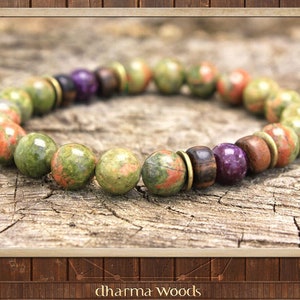 May include: A beaded bracelet featuring round unakite stones in shades of green and pink, interspersed with dark brown wooden beads, a single purple bead, and small metallic brass-toned spacers. The text "dharma Woods" is visible at the bottom.