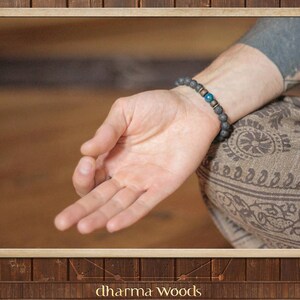 May include: A beaded bracelet featuring dark gray porous lava stone beads, two small metallic spacers, and a single smooth, round, vibrant blue bead. The bracelet is worn on a wrist, with the beads arranged in a circular pattern around the arm.