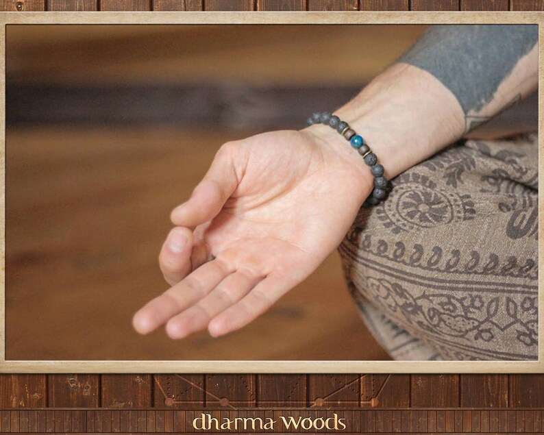 May include: A beaded bracelet featuring dark gray porous lava stone beads, two small metallic spacers, and a single smooth, round, vibrant blue bead. The bracelet is worn on a wrist, with the beads arranged in a circular pattern around the arm.
