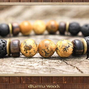 May include: A beaded bracelet featuring alternating black porous lava stone beads, smooth dark brown wooden beads, and tan speckled jasper beads. The beads are separated by thin, flat, circular brass-colored metal spacers. The text dharma woods is visible.