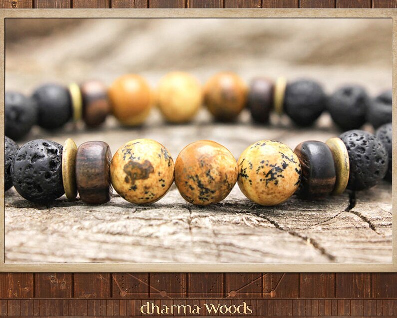 May include: A beaded bracelet featuring alternating black porous lava stone beads, smooth dark brown wooden beads, and tan speckled jasper beads. The beads are separated by thin, flat, circular brass-colored metal spacers. The text dharma woods is visible.