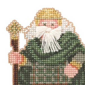 May include: A cross-stitch ornament depicting a figure in traditional Scottish attire. The figure wears a green plaid cape, brown trousers, and boots. A white beard and blonde hair are visible. The figure holds a staff and stands on a beaded base.