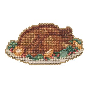 May include: A cross-stitched Thanksgiving turkey on a white plate, surrounded by green leaves, orange slices, and red berries. The turkey is brown and tan, with a detailed texture. The plate has a gold border.