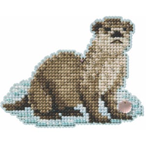 May include: A cross-stitch otter design in shades of brown and beige, with a light blue border. A small pink seashell is near the otter. The otter is lying down, with its head and body in profile.