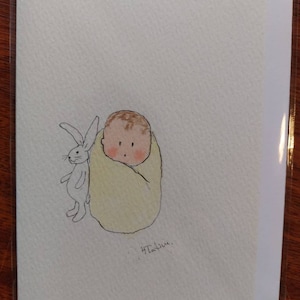 Könnte beinhalten: Eine Aquarell-Illustration eines Babys, das in eine gelbe Decke gewickelt ist, mit einem weißen Kaninchen daneben. Die Illustration befindet sich auf einer weißen Karte mit dem Wort "Ta-da!" in Kursivschrift am unteren Rand.