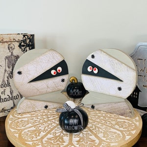 May include: Two white wooden mummy figures with black eyes and spider web details, sitting on a round wooden tray with a carved design. Three black and white Halloween-themed pumpkins with the words "Spooky", "Happy Halloween", and "Boo" are on the tray.