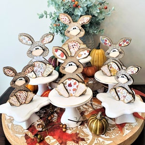 May include: Six wooden bunny figurines with fall-themed fabric patterns are displayed on white cake stands. The stands are arranged on a wooden tray with fall leaves and gourds.
