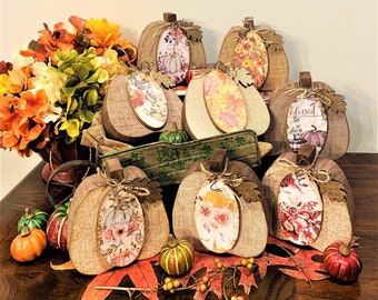 May include: A collection of eight wooden pumpkins decorated with burlap and floral designs. The pumpkins are arranged on a wooden table with fall-themed decorations, including leaves and gourds.