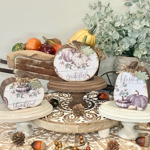 May include: Three wooden pumpkin-shaped signs featuring printed illustrations of pumpkins, cotton sprigs, and the words thankful, grateful, and blessed. Each sign is accented with a small jute twine bow and a green fabric leaf, displayed on tiered wooden stands.