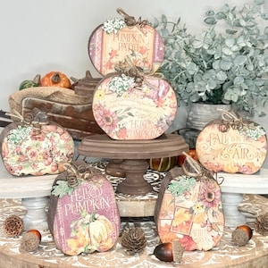 May include: A collection of five flat, pumpkin-shaped wooden decorations featuring autumn-themed floral prints, fall foliage, and text including "Hello Pumpkin" and "Fall is in the Air." Each piece has a small wire stem and a decorative paper leaf.