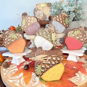 May include: A collection of decorative acorn ornaments with a wooden top and a painted bottom in various colors. Each acorn is adorned with a twine bow and a small leaf. The acorns are displayed on white pedestals and a wooden tray with fall leaves.