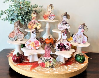 May include: A collection of eight decorative gourds with fall-themed designs, displayed on white tiered stands. The gourds are made of wood and feature various patterns and colors, including floral, geometric, and abstract designs. The gourds are arranged in a circular pattern on the stands, creating a visually appealing display.