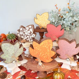 May include: Six wooden leaf decorations in various colors, including yellow, orange, pink, and green, with brown edges. Each leaf has a unique patterned design and is displayed on a small stand. A fall-themed decorative item.