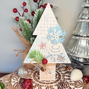 May include: A decorative wooden Christmas tree with a light blue snowflake pattern. The tree is adorned with a blue snowflake, red berries, and a small sprig of greenery. The tree is set on a wooden base.