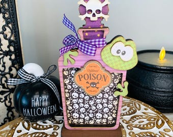 May include: A pink and purple wooden Halloween potion bottle with a skull and crossbones lid, a green frog, and a label that says "Unfiltered Poison". The bottle is decorated with a white and black skull and crossbones pattern.