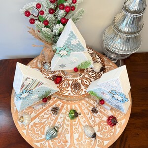 May include: A festive Christmas display featuring three white pyramid-shaped ornaments with snowflake and patterned accents. The ornaments are arranged on a decorative wooden platter with various holiday decorations, including pinecones, ornaments, and faux berries. A frosted evergreen branch and a silver tree are also present.