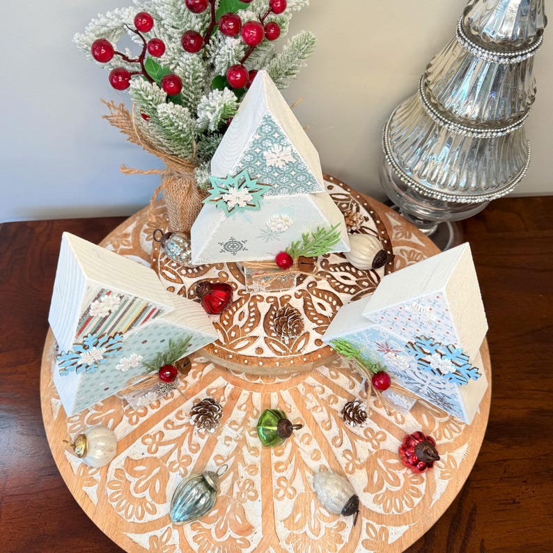 May include: A festive Christmas display featuring three white pyramid-shaped ornaments with snowflake and patterned accents. The ornaments are arranged on a decorative wooden platter with various holiday decorations, including pinecones, ornaments, and faux berries. A frosted evergreen branch and a silver tree are also present.