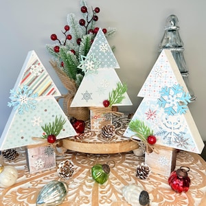 May include: Three decorative wooden Christmas trees with snowflake and greenery accents. Each tree is a different pattern, with light blue and white snowflake designs. The trees are on wooden blocks and decorated with red berries and ornaments.