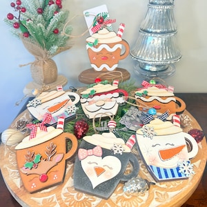 May include: A collection of wooden Christmas ornaments shaped like mugs of hot chocolate, each with a unique design. The ornaments feature festive elements like a reindeer, snowman, penguin, and gingerbread man, with whipped cream and candy cane accents.