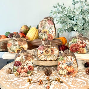 May include: A collection of five flat, wooden pumpkin-shaped decorations featuring intricate botanical prints of sunflowers, chrysanthemums, autumn leaves, and small pumpkins. Each piece is topped with a rustic twine bow and natural wood edges.