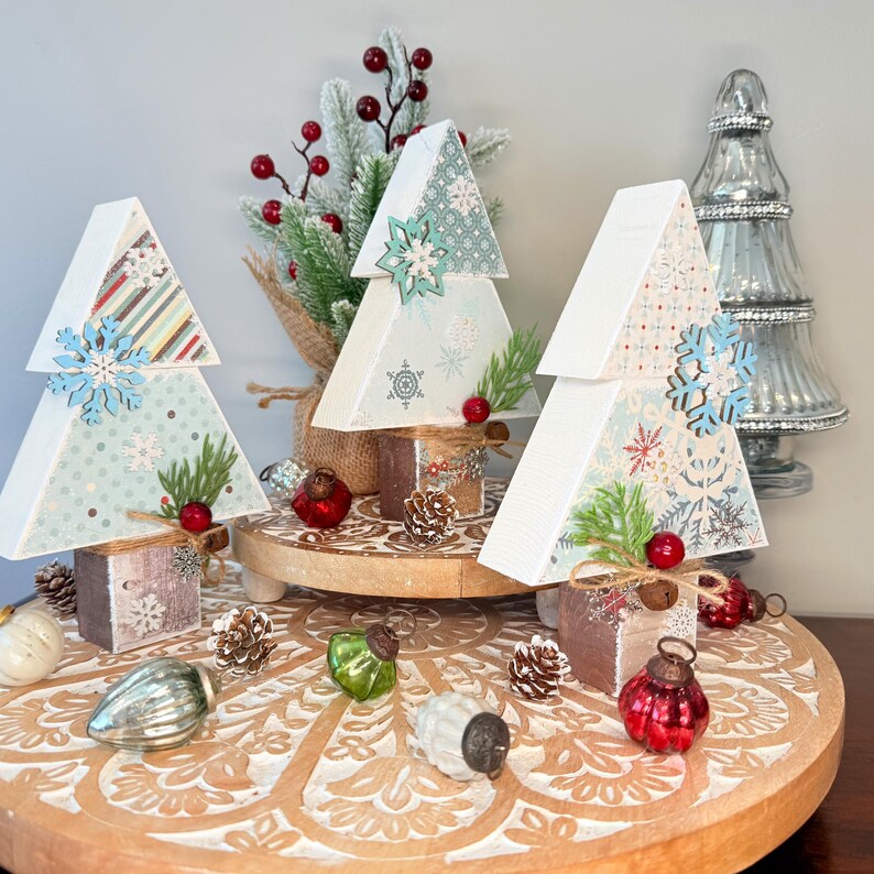 May include: Three decorative wooden Christmas trees with snowflake accents and patterned paper designs. Each tree is adorned with a wooden snowflake, faux greenery, and red berries. The trees sit on a wooden stand with vintage glass ornaments and pinecones.