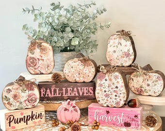 May include: A collection of wooden pumpkin-shaped decorations and rectangular blocks featuring floral patterns in shades of pink and brown. The blocks display the text "FALL LEAVES", "Pumpkin", and "harvest" alongside small pinecones and greenery.