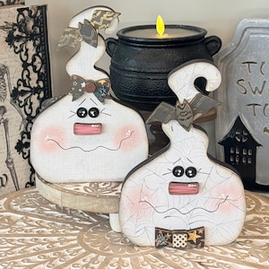 May include: Two white wooden ghost-shaped decorations with black button eyes, pink painted cheeks, and wavy mouths. One ghost features a patterned bow at the neck and a bat topper, while the other has a spider-web print and a bat on its head.